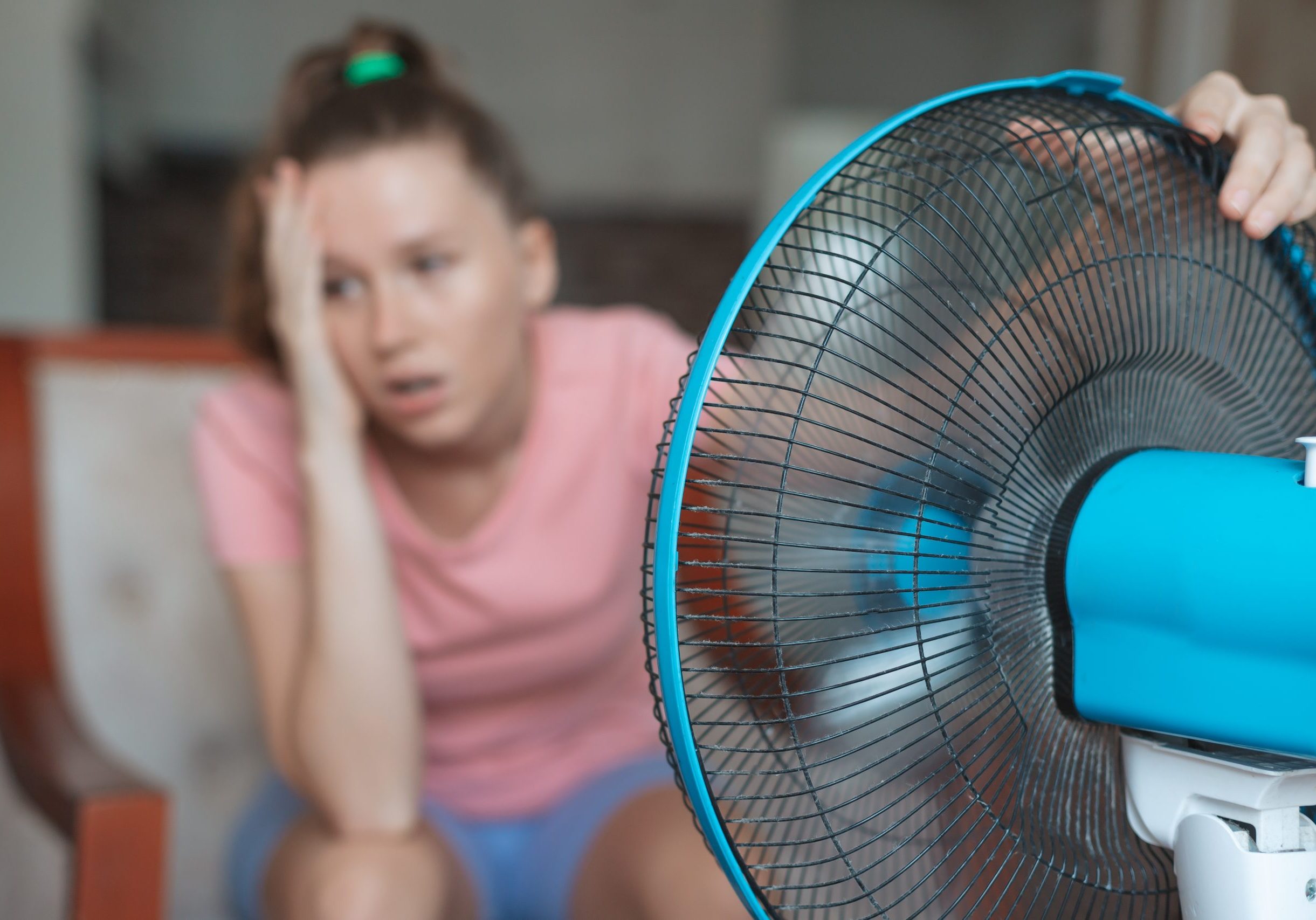 Young woman using electric fan at home in living room, sitting on couch cooling off during hot weather, suffering from heat, high temperature